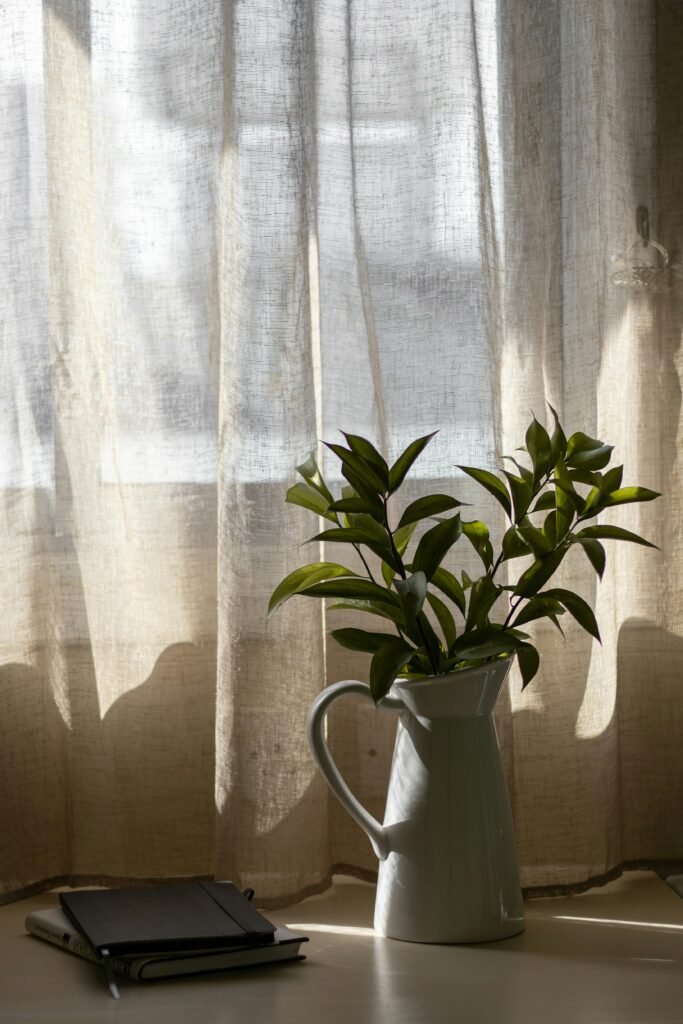 sunlight coming in through natural cottage linen curtains resting on greenery in a vase beside two journals