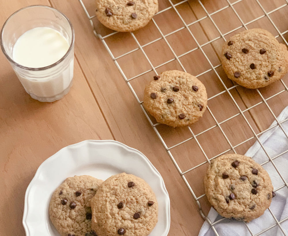 a glass of milk beside a wire cooling rack filled with chocolate chip cookies and a hand towel next to a white plate of cookies