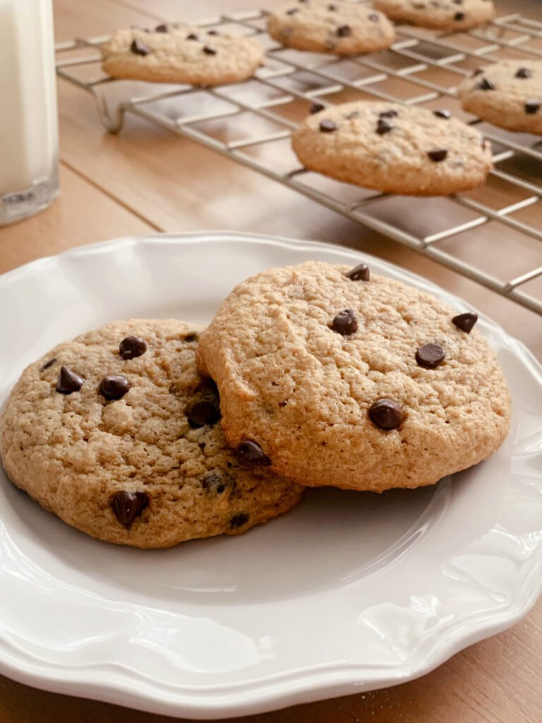 two fresh milled chocolate chip cookies on a white plate near a cooling rack filled with cookies