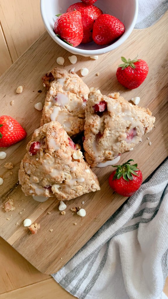 Fresh milled white chocolate strawberry scones layered on a wooden cutting board with strawberries, white chocolate chips, crumbs next to a tea towel
