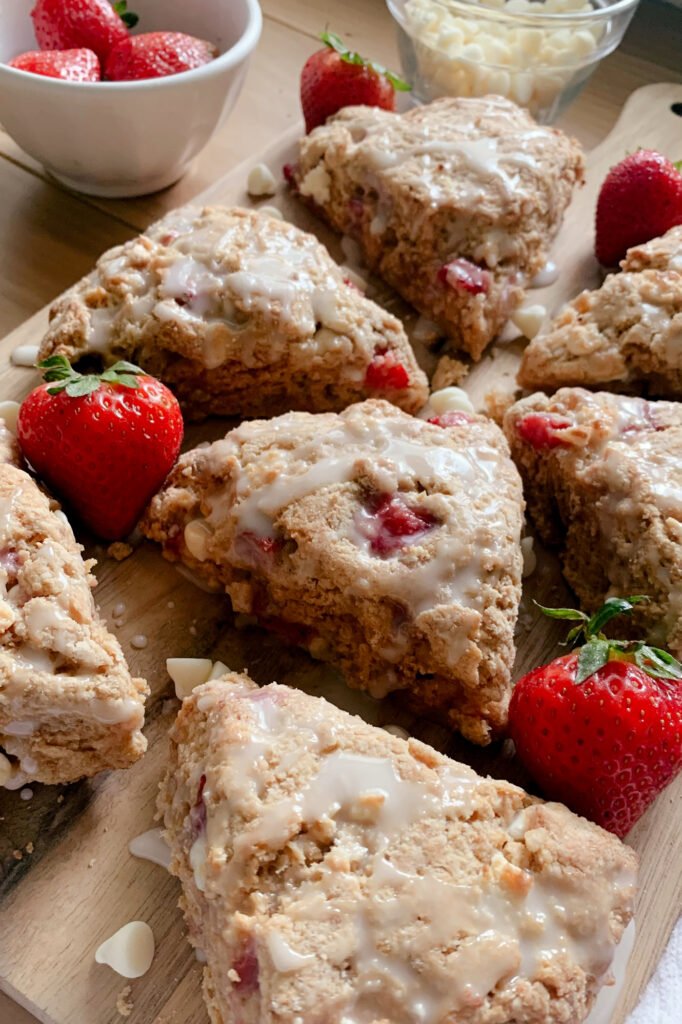 fresh milled white chocolate strawberry scones drizzled with icing on a wooden cutting board with strawberries and a bowl of white chocolate chips on a wooden table