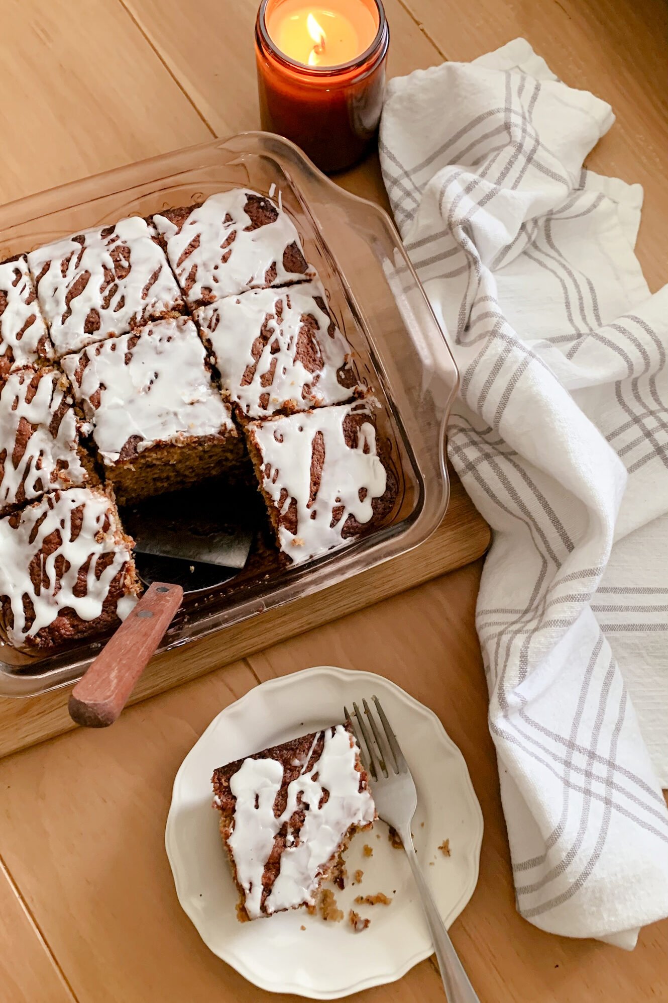Fresh milled flour banana bread coffee cake next to a lit candle, tea towel, and slice of cake on a plate