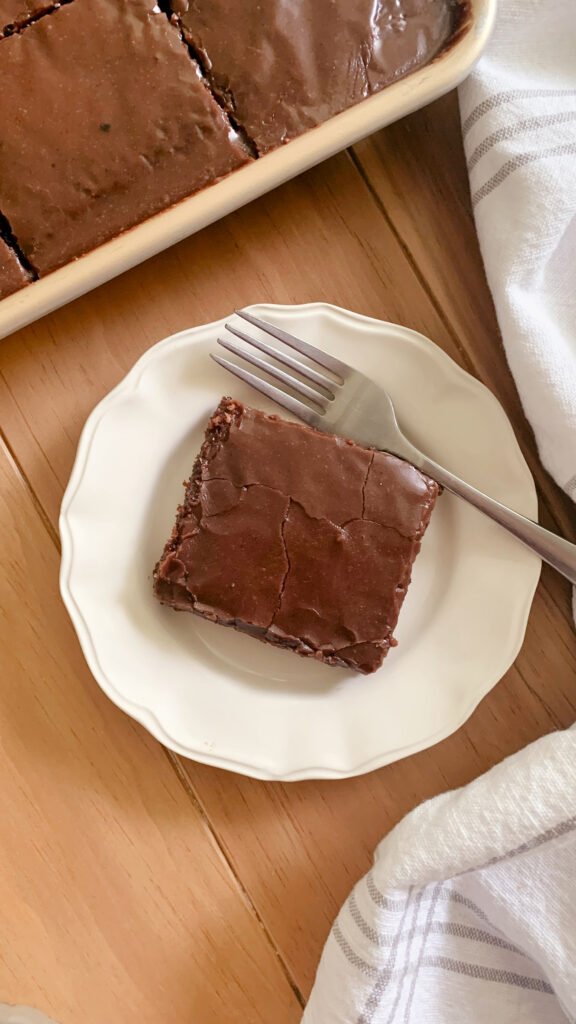 Fresh milled Texas Sheet cake cut into squares in a pan next to a tea towel and a plate with a slice of cake next to a fork on a wooden surface