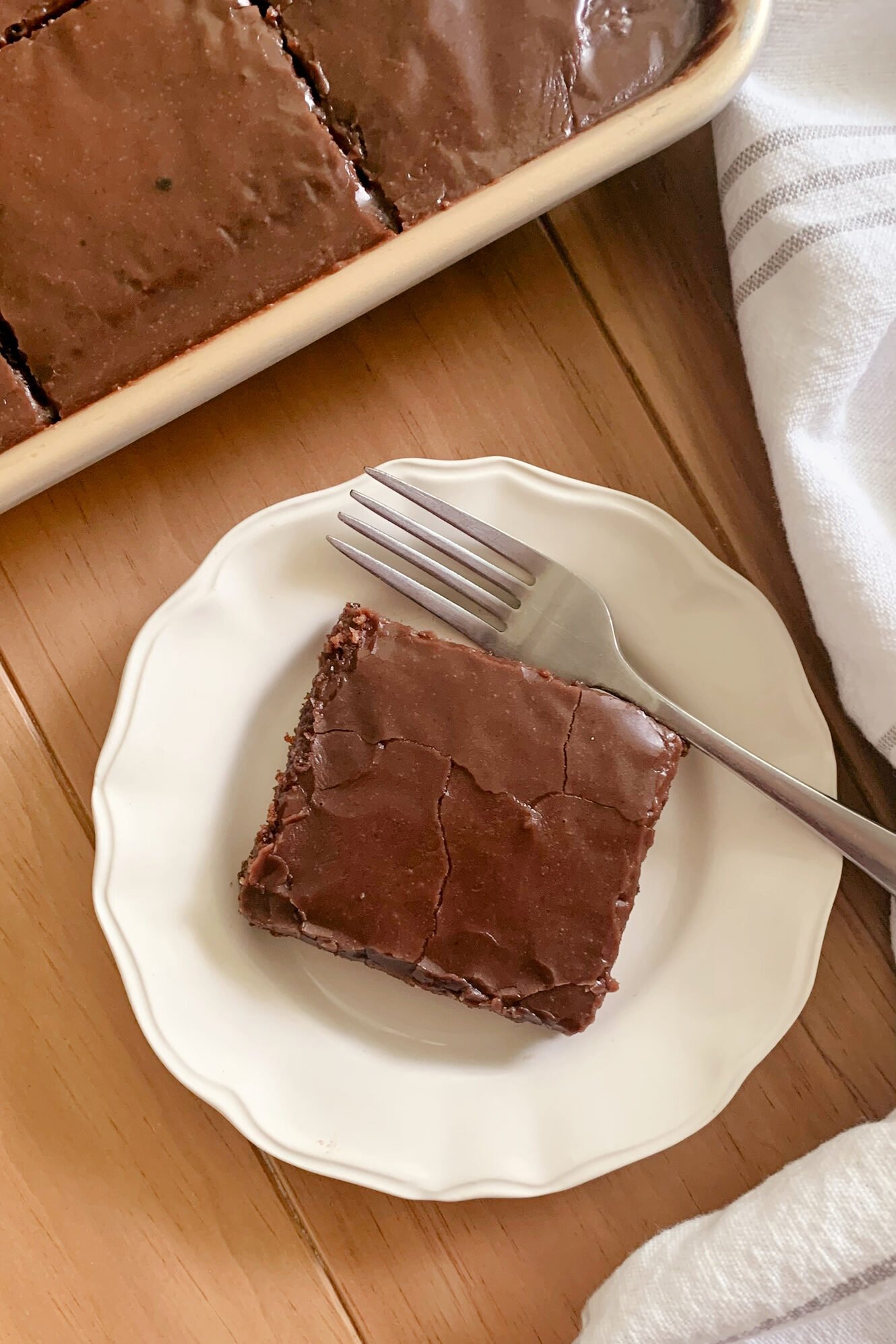 Fresh milled Texas Sheet cake cut into squares in a pan next to a tea towel and a plate with a slice of cake next to a fork on a wooden surface