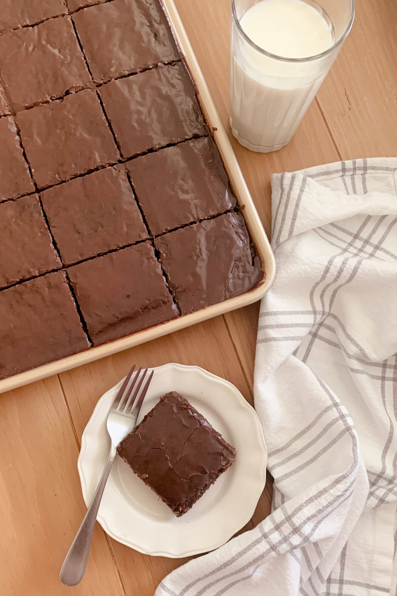 Fresh milled Texas Sheet cake cut into squares in a pan next to a glass of milk, a tea towel, and a plate with a slice of cake next to a fork on a wooden surface