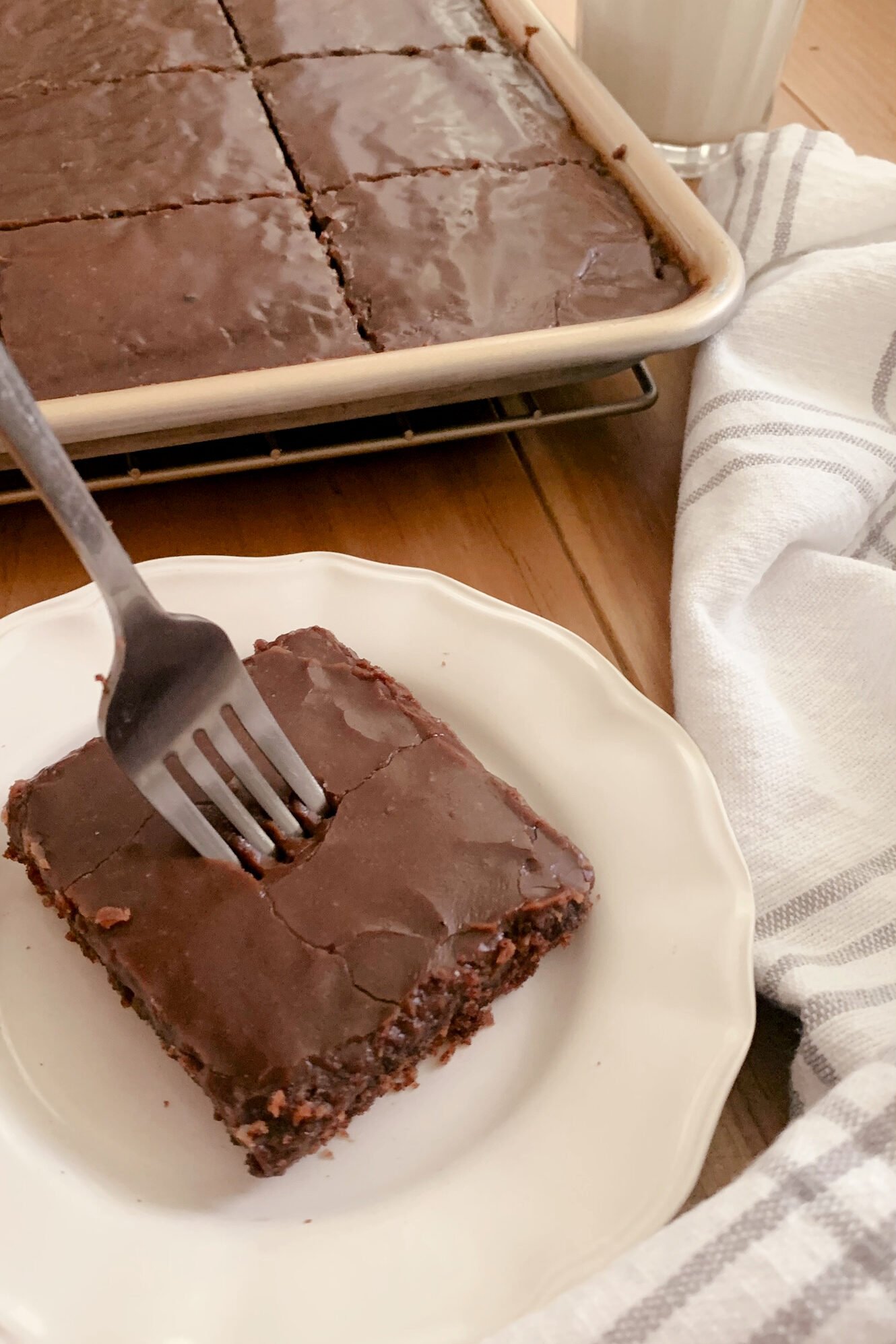 Fresh milled Texas Sheet cake cut into squares in a pan next to a glass of milk, a tea towel, and a plate with a slice of cake with a fork in it on a wooden surface