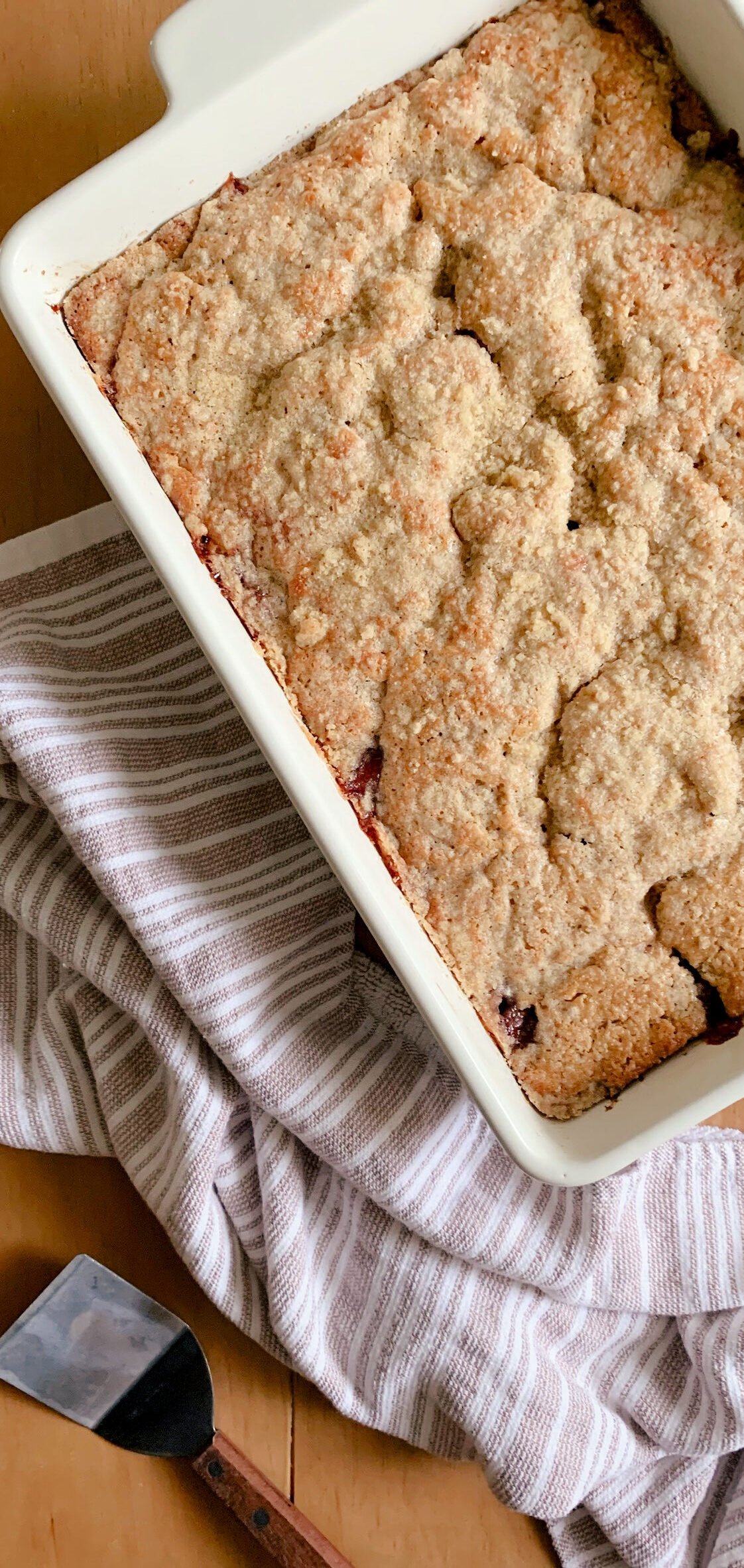 Fresh Milled Cherry Coffee Cake next to a tea towel and serving utensil on a wooden surface.