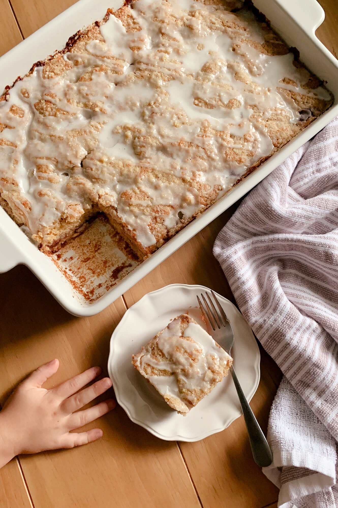 Fresh Milled Cherry Coffee Cake next to a tea towel and plate with a slice and fork, with a child's hand reaching for it.