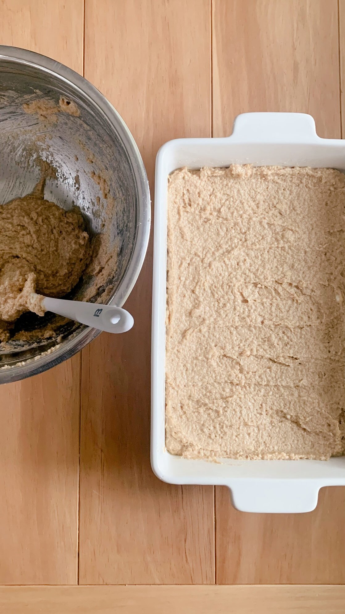 The bottom layer of fresh milled cherry coffee cake batter spread out evenly next to a stainless steel bowl of batter and a spatula, on a wooden surface