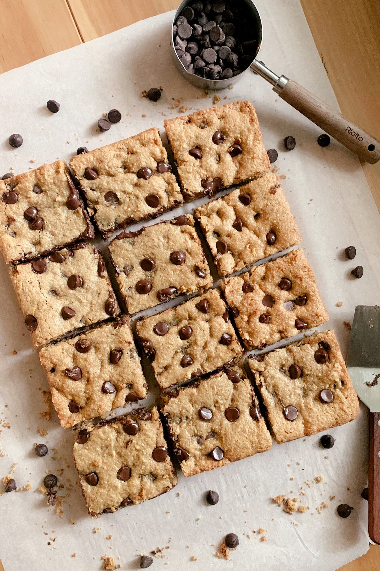 fresh milled chocolate chip cookie bars cut into squares on a piece of parchment paper next to a measuring cup of chocolate chips and a serving utensil