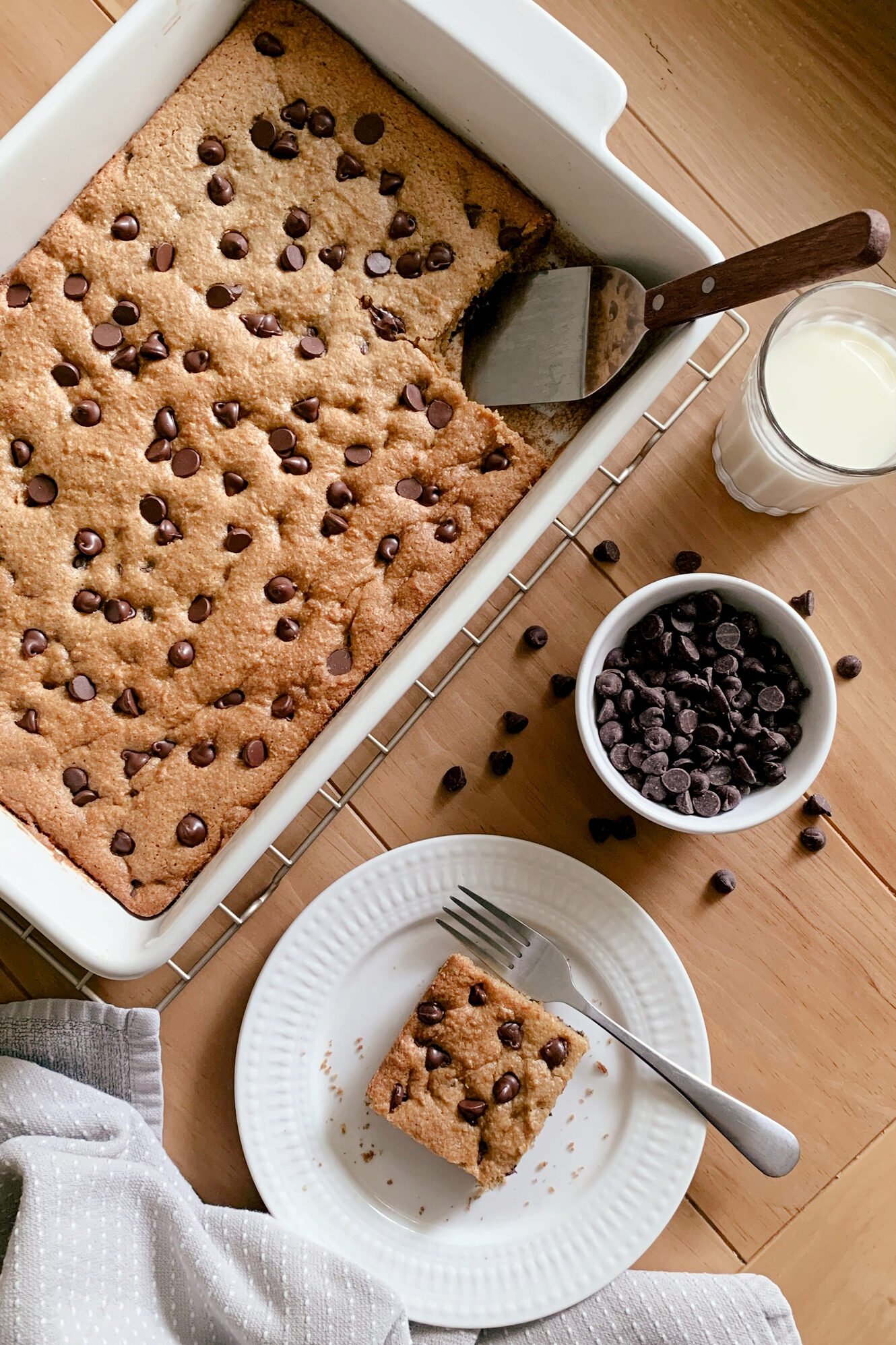 Fresh milled chocolate chip cookie bars in a 9x13 baking dish on a wooden table next to a glass of milk, a plate of a cookie bar, and a bowl of chocolate chips
