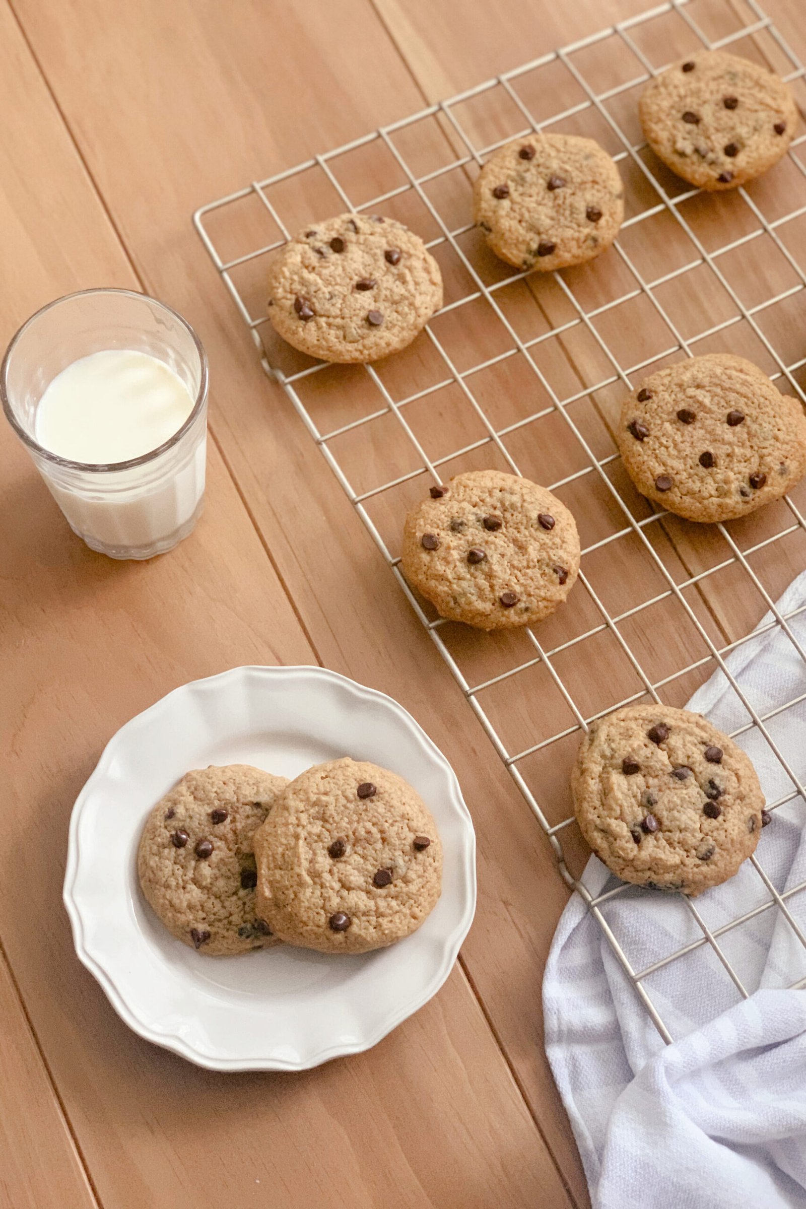a glass of milk beside a wire cooling rack filled with chocolate chip cookies and a hand towel next to a white plate of cookies