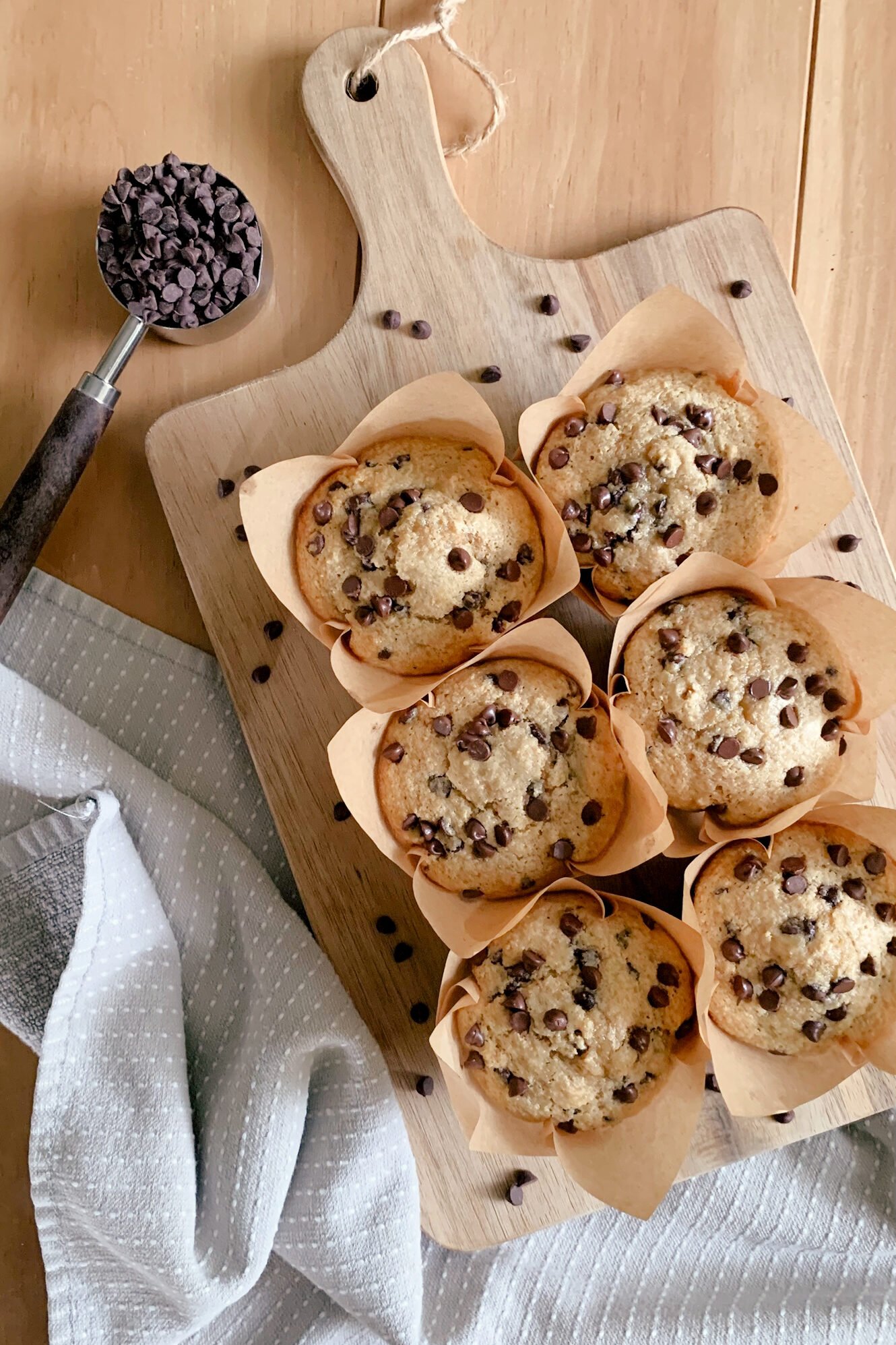 fresh milled chocolate chip muffins on a cutting board with a tea towel underneath next to a measuring cup of chocolate chips