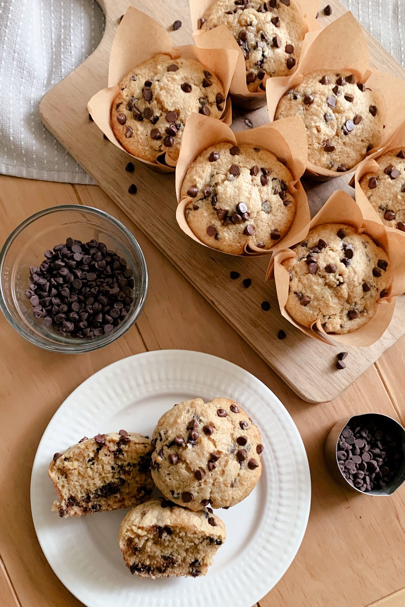 fresh milled chocolate chip muffins on a wooden cutting board with a tea towel underneath next to a bowl of chocolate chips and a plate of two more muffins