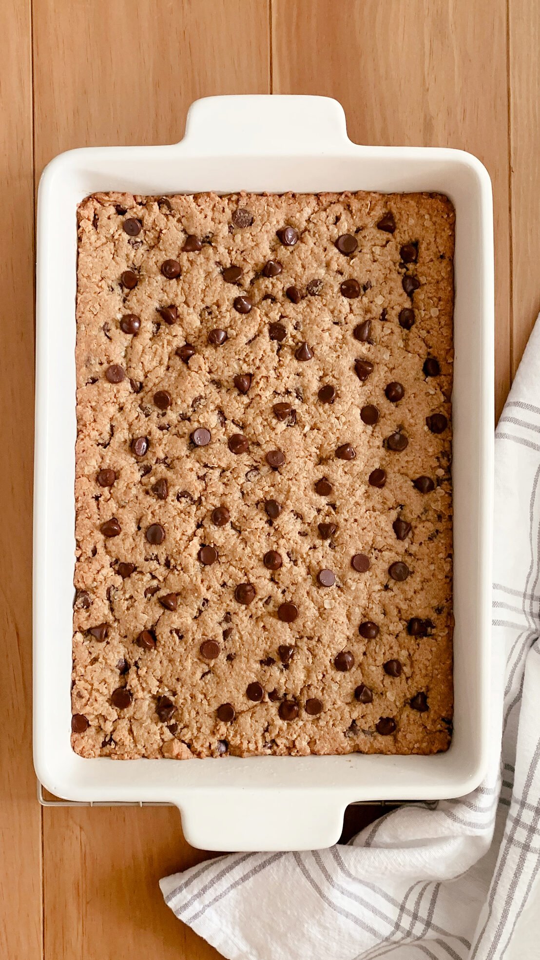 The fresh milled peanut butter oatmeal chocolate chip cookie bars in a baking dish laid out on a wooden table next to a tea towel